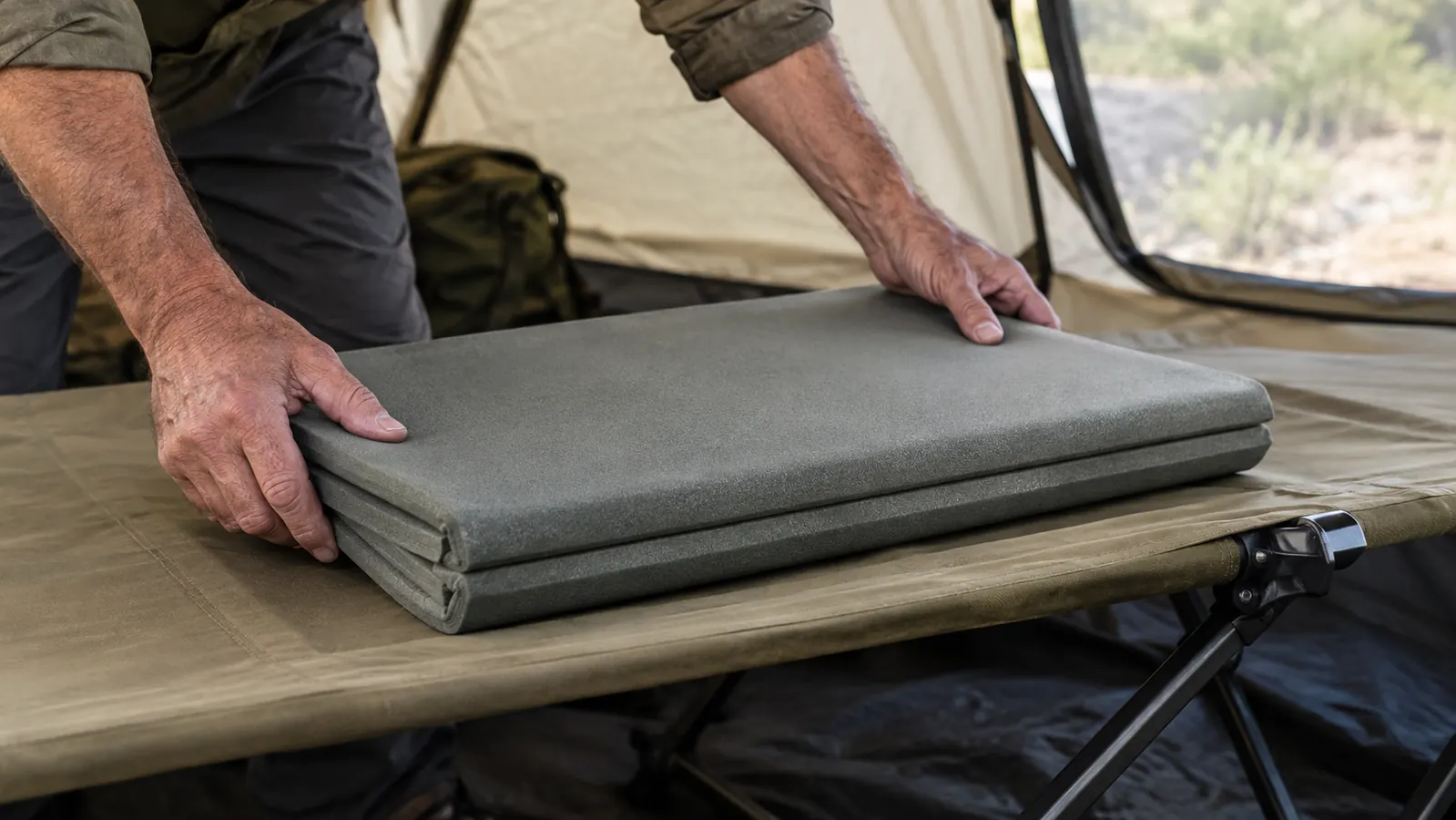 Hands laying a folded closed-cell foam pad onto the taut fabric of a camp cot, the insulation step of a cold-weather setup