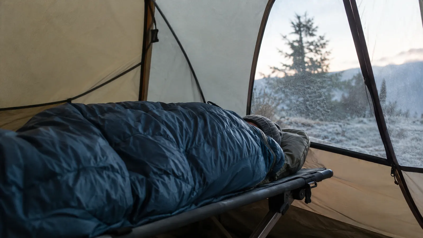 A camper asleep on a camp cot inside a tent on a cold morning, zipped into a sleeping bag, dry tent walls