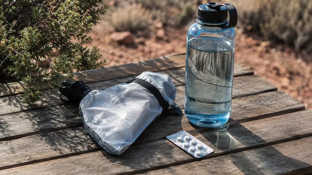 A water filter pouch and a small foil strip of tablets laid on a camp table beside a filled water bottle