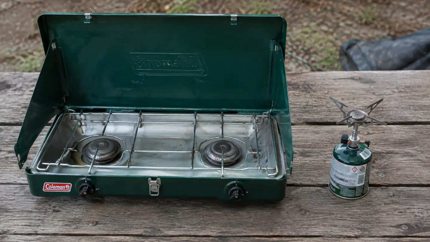 A large two-burner camp stove beside a tiny single-burner backpacking stove on a picnic table